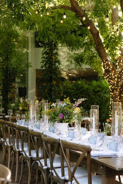 A long wooden dining table set for a wedding reception with floral centerpieces, glassware, and string lights hanging overhead.