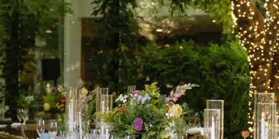 A long wooden dining table set for a wedding reception with floral centerpieces, glassware, and string lights hanging overhead.