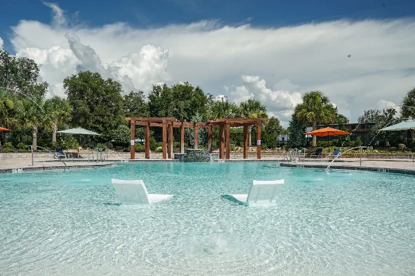 A large outdoor swimming pool with white lounge chairs, surrounded by palm trees and a wooden pergola structure under a cloudy sky.