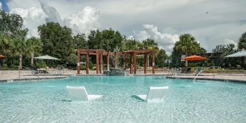 A large outdoor swimming pool with white lounge chairs, surrounded by palm trees and a wooden pergola structure under a cloudy sky.