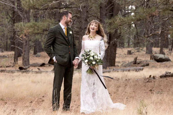 A bride and groom standing in a pine forest, the bride holding a bouquet of white flowers.