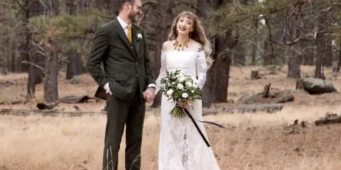 A bride and groom standing in a pine forest, the bride holding a bouquet of white flowers.
