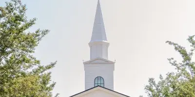 Exterior view of a white wedding chapel with a tall steeple, surrounded by trees and a paved walkway.