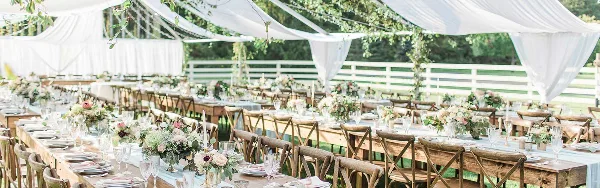An outdoor wedding reception setup featuring long wooden tables, chairs, and a large white tent.
