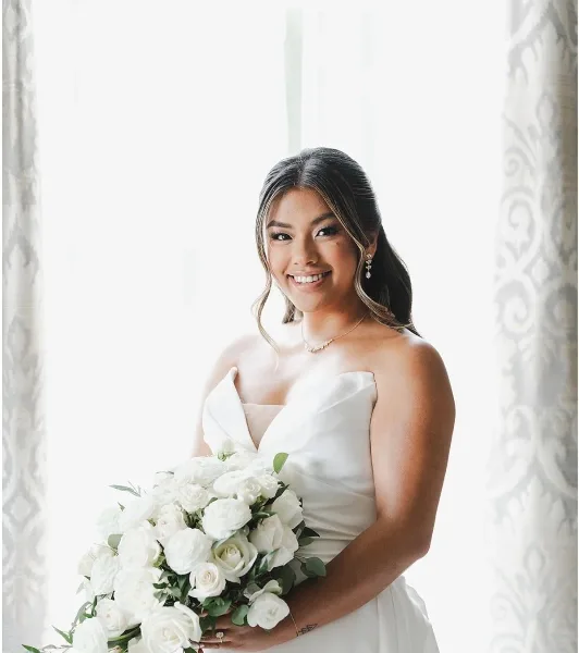 A smiling bride with styled hair and makeup holding a white rose bouquet.