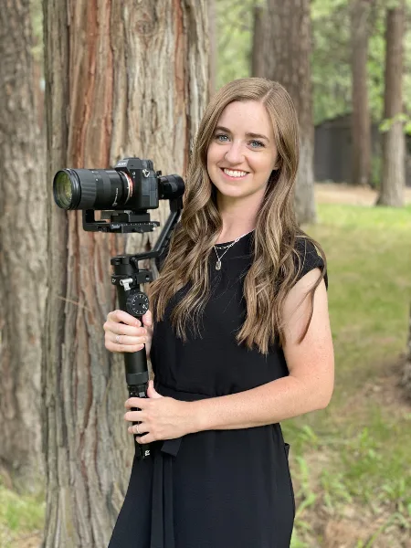A woman in a black dress holding a professional video camera on a gimbal in a forest setting.