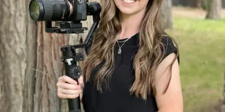 A woman in a black dress holding a professional video camera on a gimbal in a forest setting.