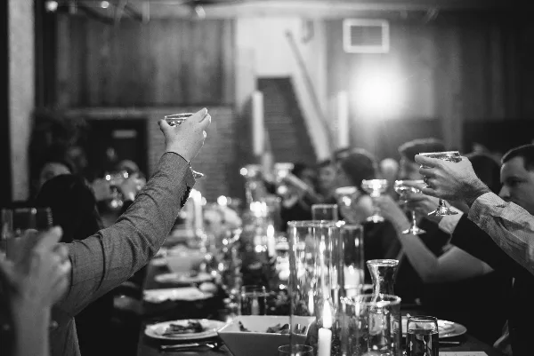 Black-and-white photo of wedding guests toasting with champagne glasses at a candlelit reception table.