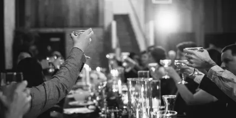 Black-and-white photo of wedding guests toasting with champagne glasses at a candlelit reception table.