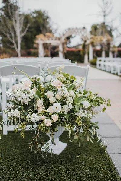 A lush white and blush floral arrangement in a white urn placed in a wedding aisle with white chairs and a floral arch in the background.