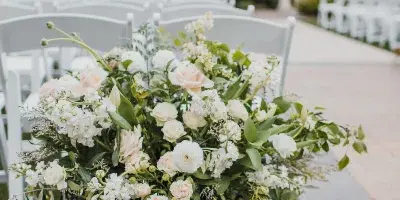 A lush white and blush floral arrangement in a white urn placed in a wedding aisle with white chairs and a floral arch in the background.