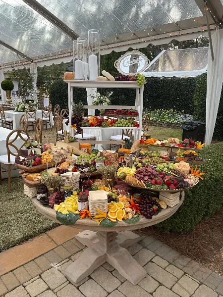 An outdoor catering table under a tent featuring a tiered display of fresh fruits, cheeses, and meats.