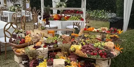 An outdoor catering table under a tent featuring a tiered display of fresh fruits, cheeses, and meats.