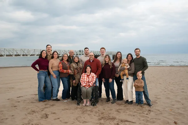 A large multi-generational family posing for a professional portrait on a sandy beach with a pier in the background.