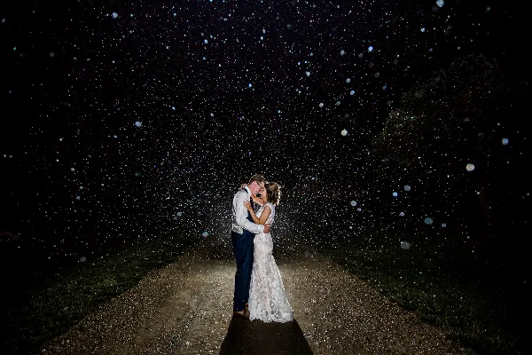 A bride and groom embracing on a dirt path at night with sparkling bokeh lights in the background.