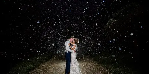 A bride and groom embracing on a dirt path at night with sparkling bokeh lights in the background.