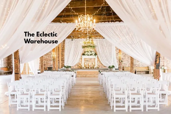 A wedding ceremony setup in a rustic indoor venue featuring white chairs, a floral altar, and hanging chandeliers.