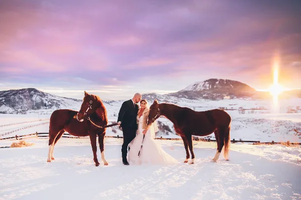 A bride and groom standing in a snowy field with two brown horses, with mountains and a sunset in the background.