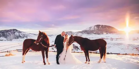 A bride and groom standing in a snowy field with two brown horses, with mountains and a sunset in the background.