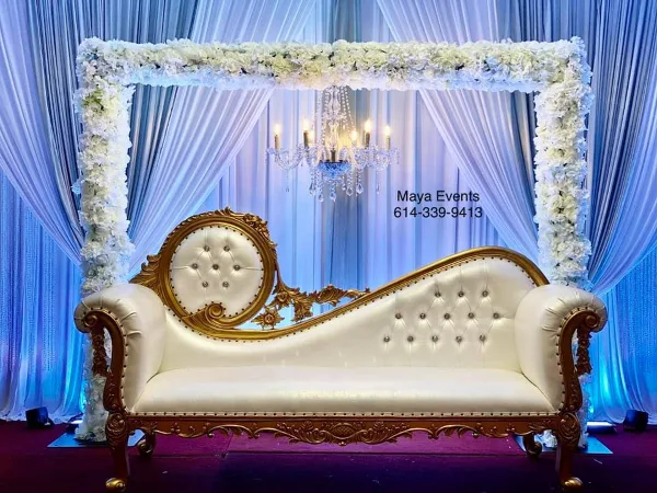 A white and gold ornate loveseat positioned under a floral arch with blue drapery and a chandelier, representing an event decoration setup.