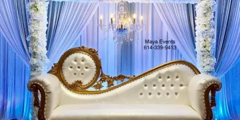 A white and gold ornate loveseat positioned under a floral arch with blue drapery and a chandelier, representing an event decoration setup.