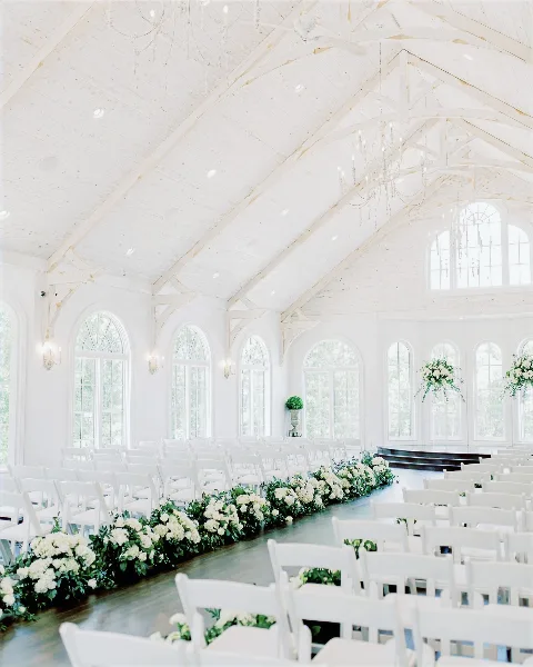 White wedding ceremony venue interior with rows of white chairs, floral aisle decorations, chandeliers, and tall arched windows.