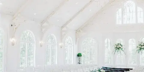 White wedding ceremony venue interior with rows of white chairs, floral aisle decorations, chandeliers, and tall arched windows.