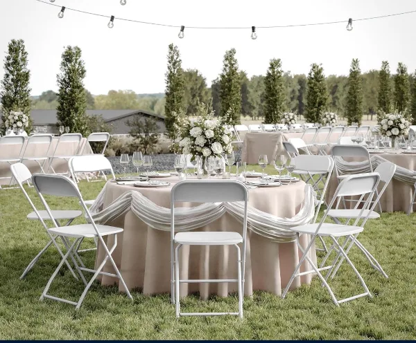 Outdoor event setup with white folding chairs, round tables with beige linens, and floral centerpieces on a lawn.