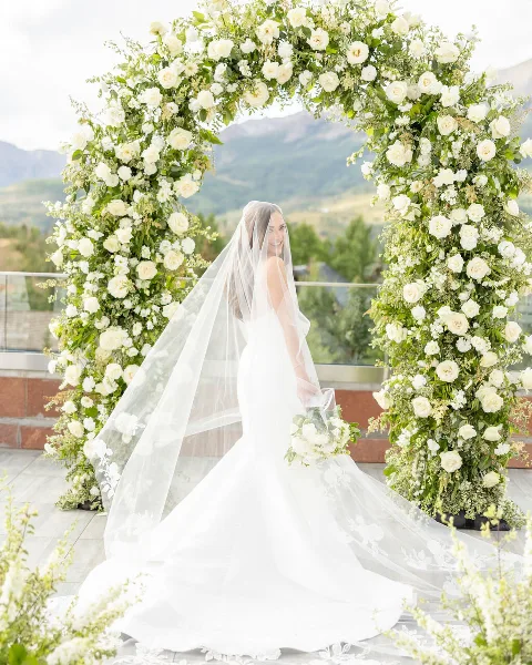 A bride in a white wedding dress and veil stands smiling beneath a large floral arch with mountains in the background.