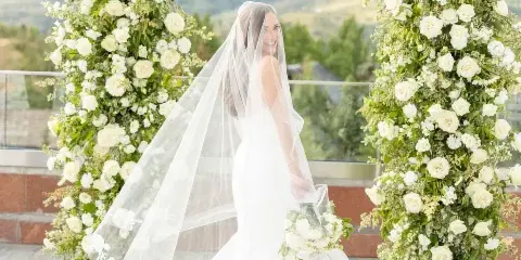 A bride in a white wedding dress and veil stands smiling beneath a large floral arch with mountains in the background.