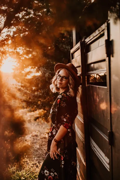 A professional portrait of a woman with blonde hair, glasses, and a brown hat, leaning against a wooden gate in a sunlit, rustic outdoor setting.
