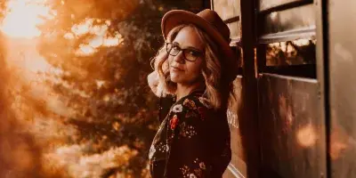 A professional portrait of a woman with blonde hair, glasses, and a brown hat, leaning against a wooden gate in a sunlit, rustic outdoor setting.