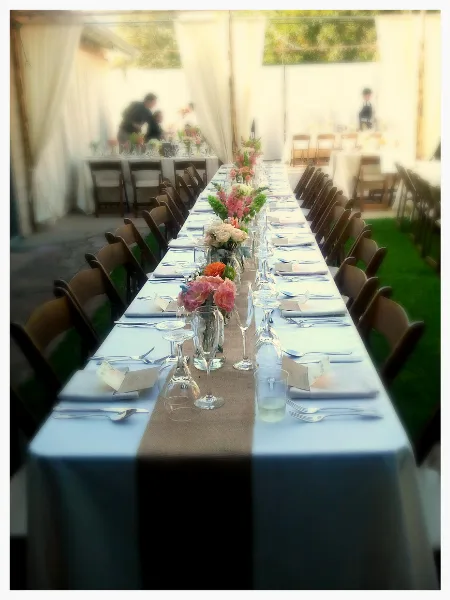 A long banquet table set with white linens, glassware, silverware, and floral centerpieces at an outdoor event venue.