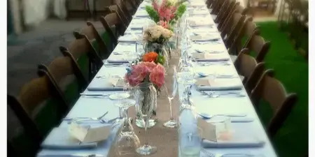 A long banquet table set with white linens, glassware, silverware, and floral centerpieces at an outdoor event venue.