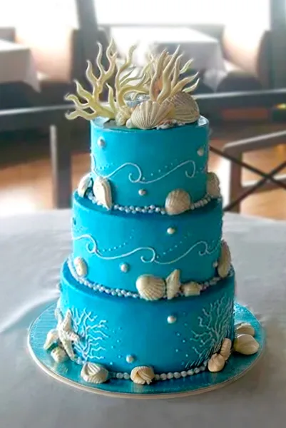 A three-tiered turquoise wedding cake decorated with white fondant shells, coral, and pearl accents.