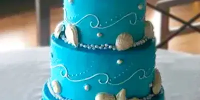 A three-tiered turquoise wedding cake decorated with white fondant shells, coral, and pearl accents.