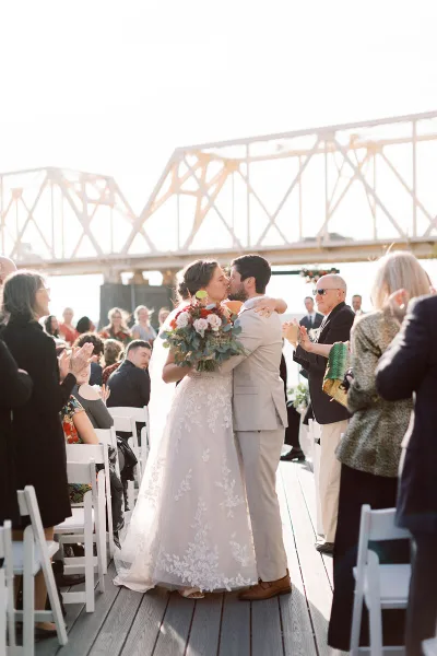 A bride and groom kiss during an outdoor wedding ceremony on a wooden deck with a bridge in the background, surrounded by applauding guests.