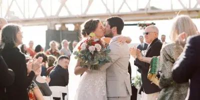 A bride and groom kiss during an outdoor wedding ceremony on a wooden deck with a bridge in the background, surrounded by applauding guests.