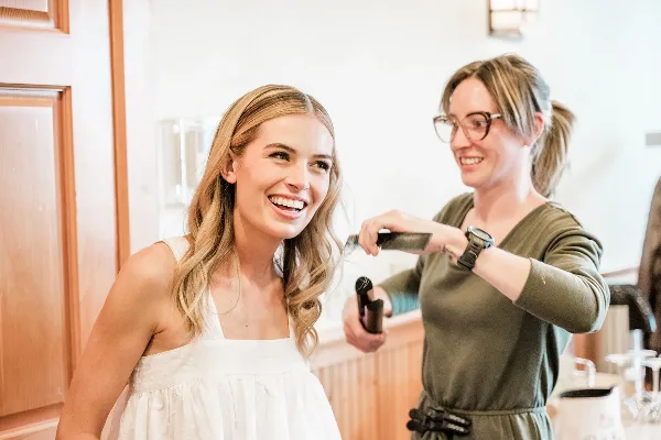 A smiling bride in a white dress receiving hair styling from a professional artist using a curling iron.