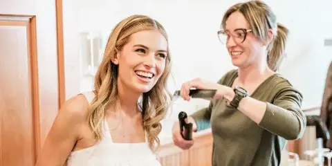 A smiling bride in a white dress receiving hair styling from a professional artist using a curling iron.