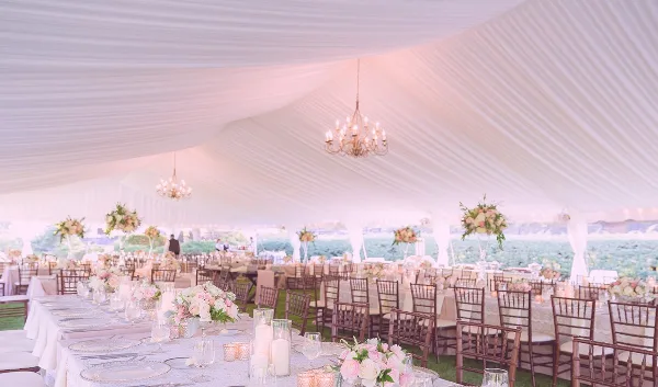 Interior of a white wedding tent with chandeliers, tables set with linens and floral centerpieces, and wooden chairs.