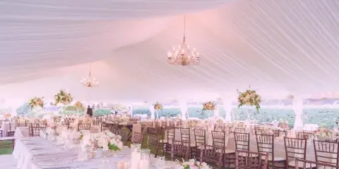 Interior of a white wedding tent with chandeliers, tables set with linens and floral centerpieces, and wooden chairs.