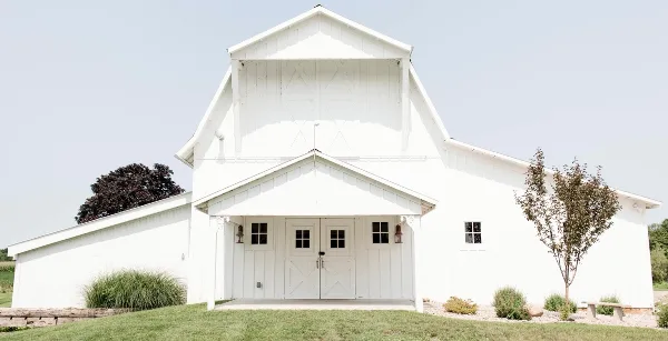 Exterior view of a white historic barn and farmhouse wedding venue with a front porch, set on a grassy lawn.