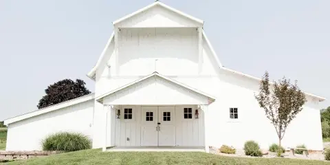 Exterior view of a white historic barn and farmhouse wedding venue with a front porch, set on a grassy lawn.