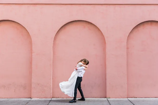 A bride and groom embracing in front of a pink arched wall.