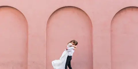 A bride and groom embracing in front of a pink arched wall.