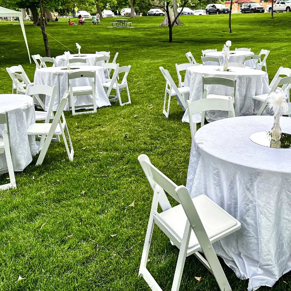 Outdoor event setup with white round tables, white folding chairs, and floral centerpieces on a grassy lawn.