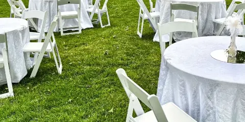 Outdoor event setup with white round tables, white folding chairs, and floral centerpieces on a grassy lawn.