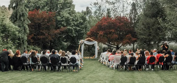 An outdoor wedding ceremony in progress at a garden venue, featuring a floral arch and rows of seated guests.