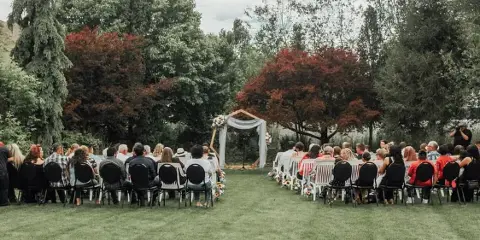 An outdoor wedding ceremony in progress at a garden venue, featuring a floral arch and rows of seated guests.
